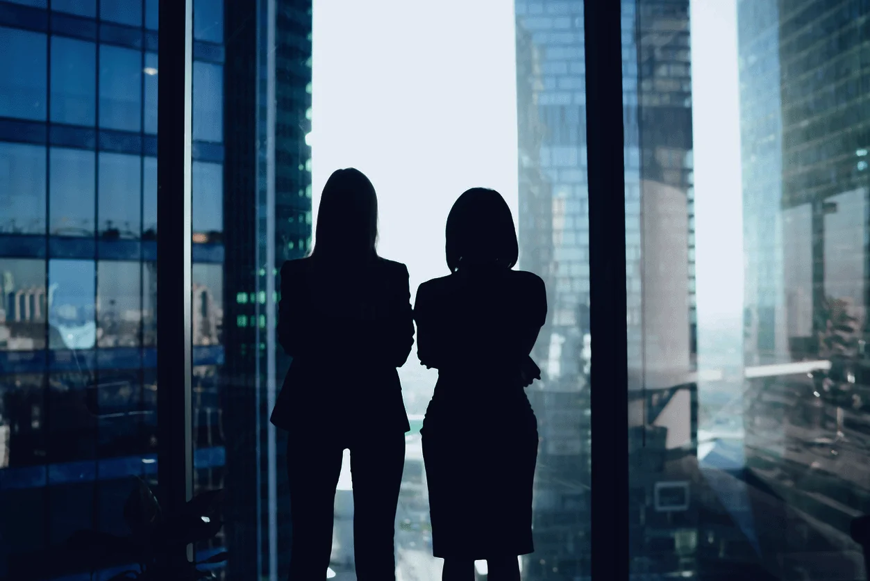 Back view of female colleagues in formal wear standing near window looking at modern exterior of skyscrapers in business center, silhouette of women together planning future success of brainstorming